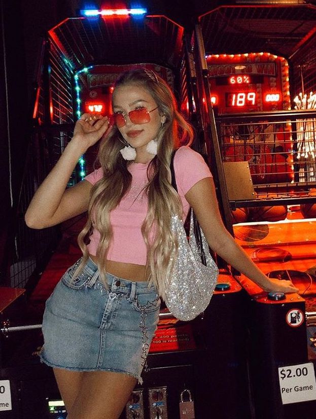 A woman channels early 2000s fashion at an arcade, posing with a hand on her red heart-shaped sunglasses. She wears a pink crop top, a high-waisted denim mini skirt with a chain belt, and a sparkling silver shoulder bag.