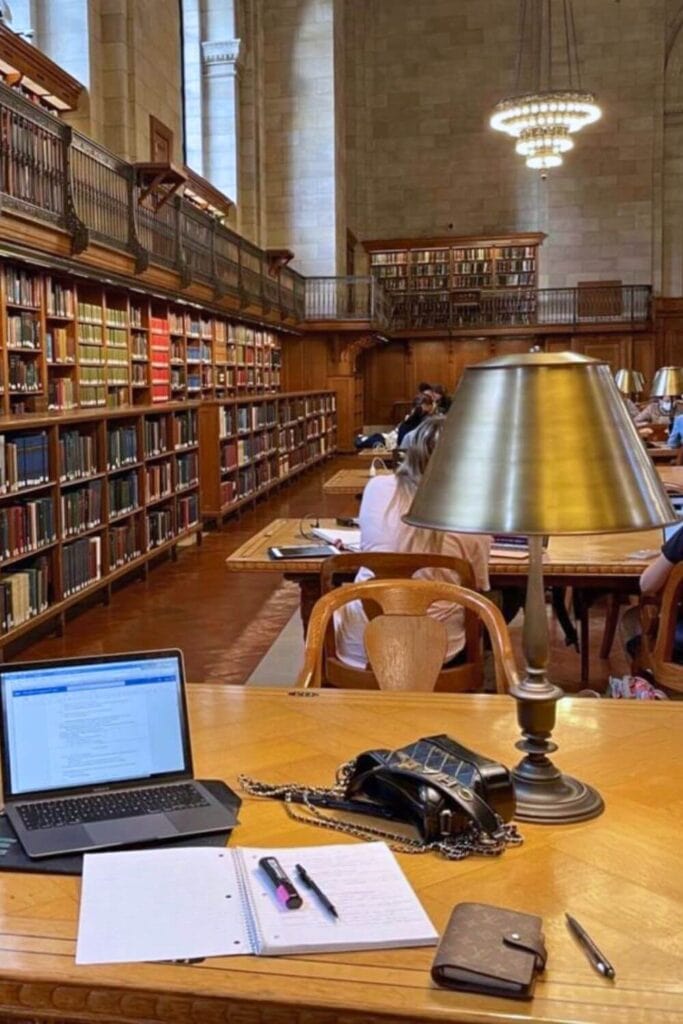 Focused students study in a grand library with high ceilings and book-lined walls; the foreground shows a study desk with an open laptop, a notebook with a highlighter, a pen, and a designer wallet.