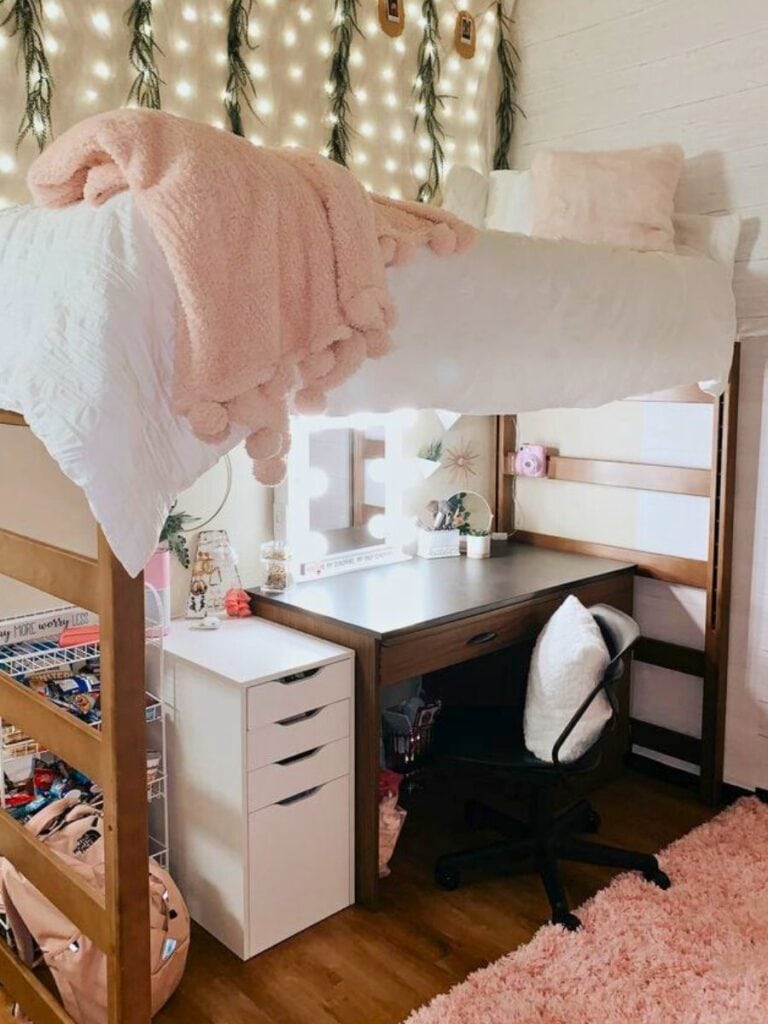 A snug lofted dorm bed with plush pink throw blankets and white pillows, under a string of fairy lights draped along a wooden-paneled wall. Below the bed is a study area with a wooden desk, white drawers, and a cozy chair, beside a shaggy pink rug that complements the room's warm and inviting aesthetic.