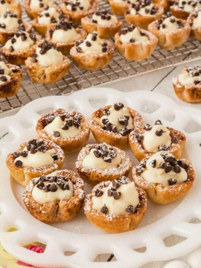 An array of mini cannoli cups sprinkled with chocolate chips and dusted with powdered sugar, elegantly presented on a white latticed tray, ideal for a celebratory dessert table.