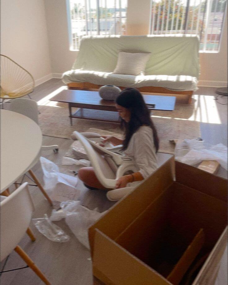 A person sits on the floor assembling furniture, surrounded by packing materials and open boxes, in a bright living room with a large window, indicating a moving in process.