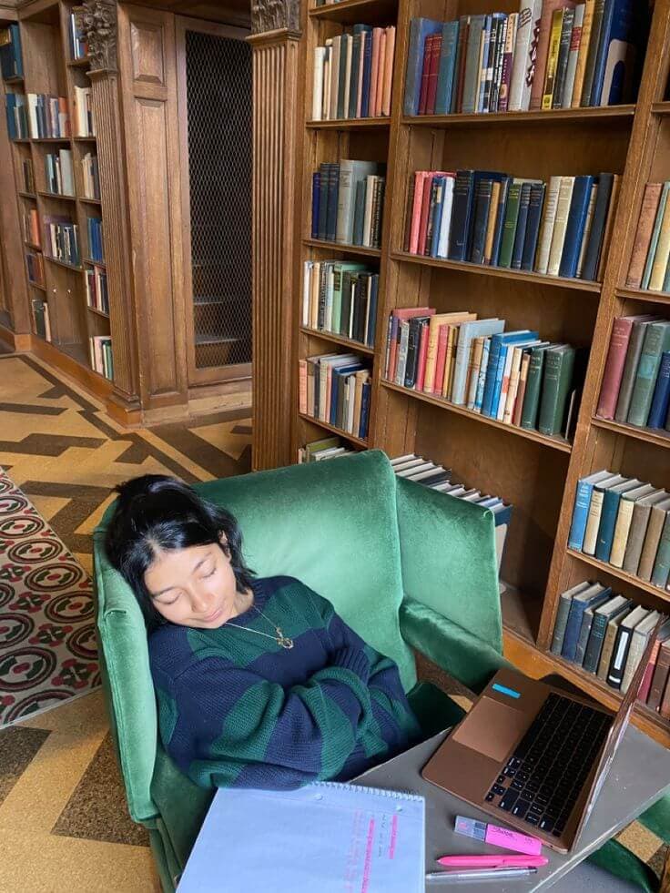 A student takes a peaceful nap in a green armchair in a quiet library corner, surrounded by bookshelves, with an open laptop and study materials on a nearby table.