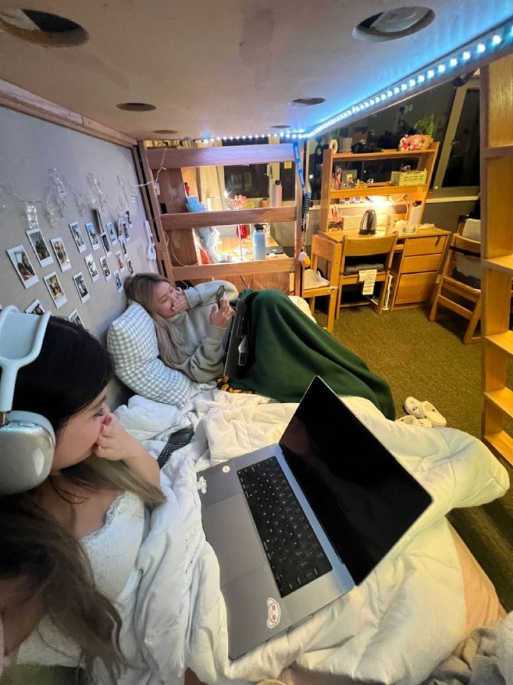 Two students relax in a cozy dorm room loft bed, with one browsing a tablet and the other wearing headphones and using a laptop, surrounded by string lights, personal photos, and a view of the room below.