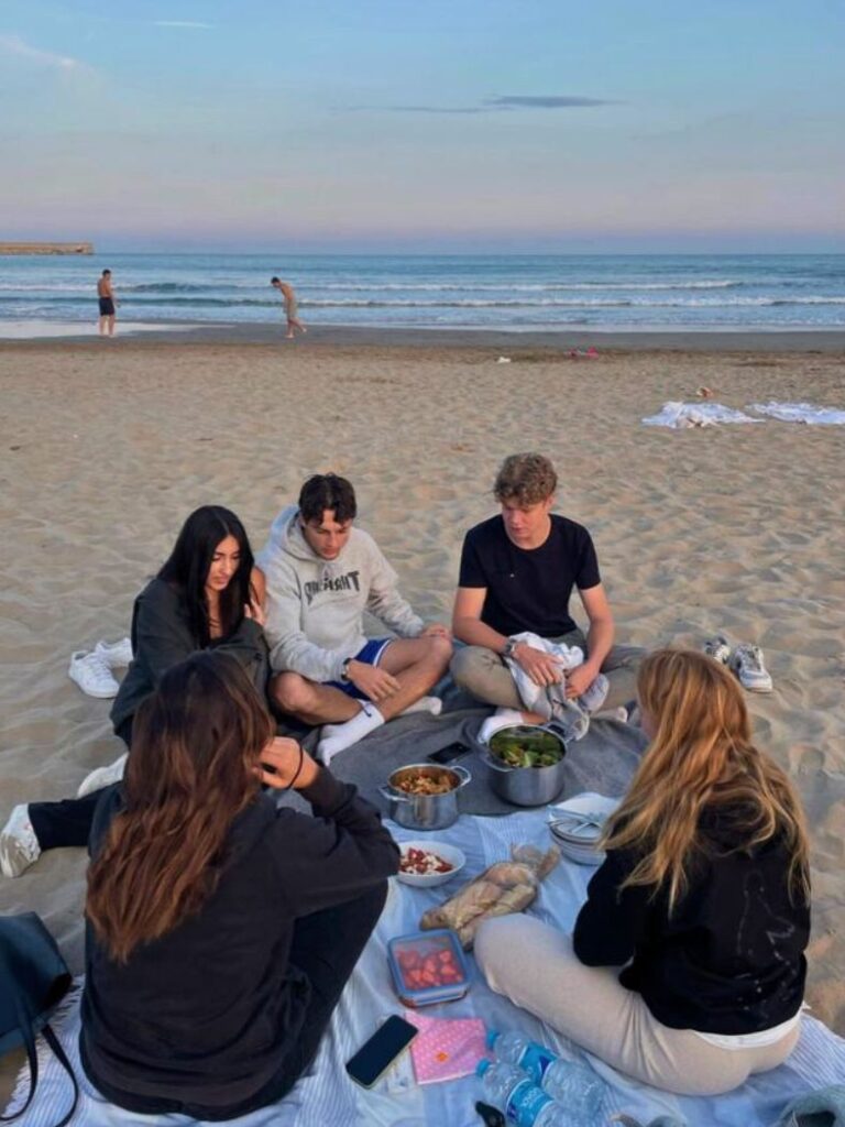 A group of young adults enjoying a beach picnic at twilight. They are seated on a blanket with a spread of food in front of them, including bowls of salad and snacks.