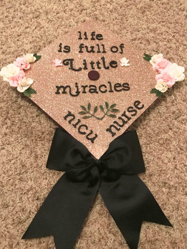 A graduation cap decoration with a glittery rose gold background and a black bow at the base. The cap features the inspiring quote "life is full of little miracles" in black script, with "NICU nurse" below in green. It's embellished with small artificial flowers in the corners, celebrating the achievement of a new nurse specializing in neonatal care.