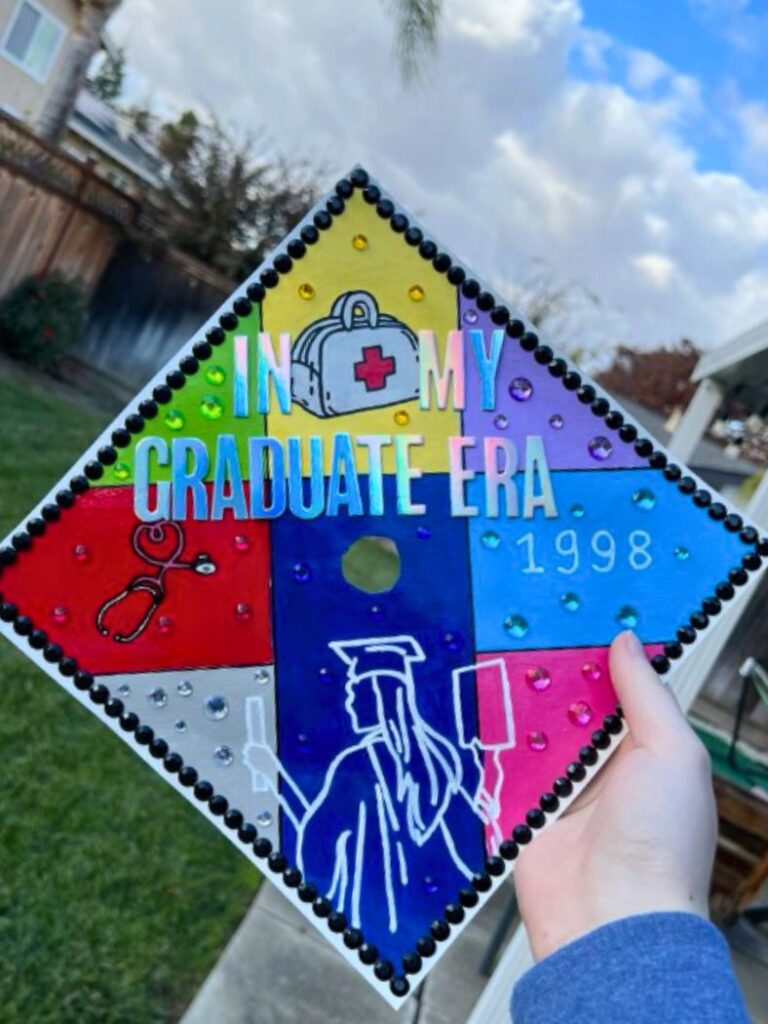 A colorfully painted graduation cap divided into quadrants with a medical theme. The cap features the statement 'IN MY GRADUATE ERA' with each quadrant showcasing a different symbol: a red cross, a stethoscope, a graduating student silhouette, and the year '1998' in blue