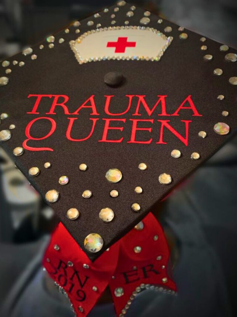 A black graduation cap with 'TRAUMA QUEEN' in bold red letters, adorned with a nurse's cap illustration with a red cross and surrounded by a scatter of iridescent rhinestones.