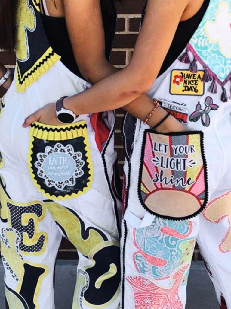 Close-up of two people wearing intricately decorated white overalls. Details include vibrant patches with inspirational messages such as 'FAITH', 'Have a nice day', and 'Let your light shine'.