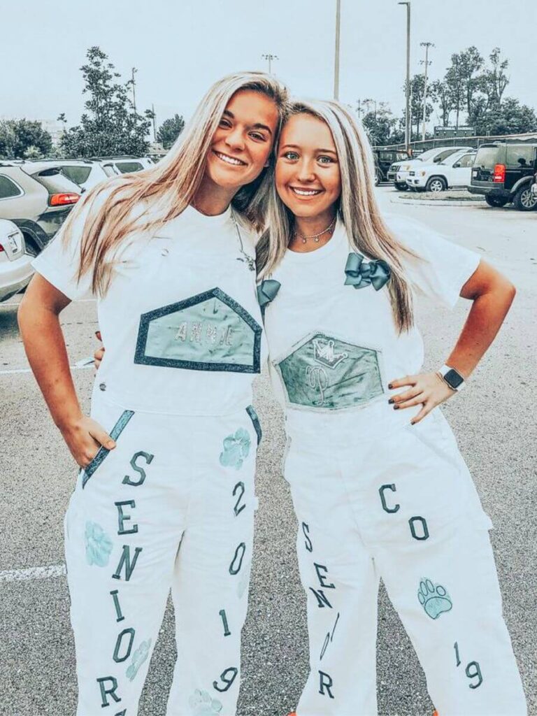 Two smiling young women wearing customized white overalls with 'SENIOR' and '2019' painted on, along with various other designs like a paw print and ribbons.